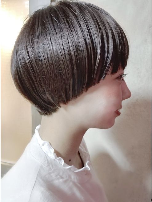 a side view portrait of a young woman with short, choppy, dark brown bob haircut with bangs. she is wearing a white ruffled shirt and has a delicate, feminine look.  the background is a plain white wall.