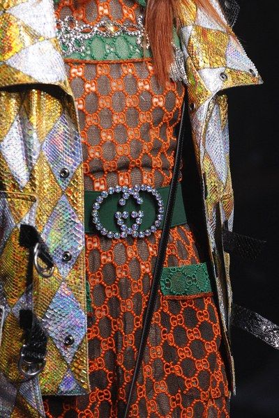 closeup shot of a woman's outfit featuring a vibrant orange gucci dress with a distinctive gg pattern, layered over a shimmering iridescent jacket with a diamond pattern. the outfit is accessorized with a gucci belt studded with crystals, and a black handbag strap is visible. the dress features a green band just above the hemline. the overall aesthetic is luxurious and highfashion, showcasing intricate details and textures. the image is likely a still from a fashion show or a highfashion editorial shoot.