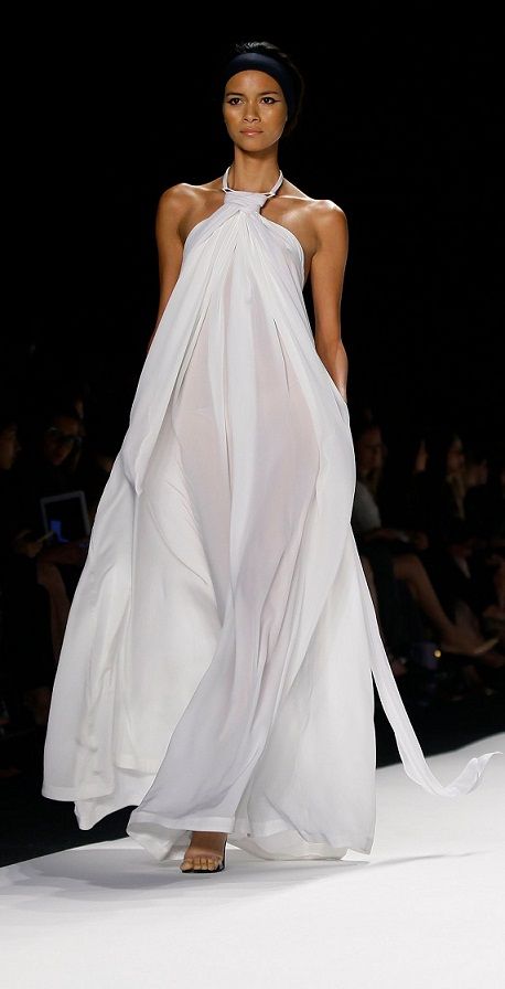 a fashion model walks the runway in a beautiful white dress, with a black headband, her hair pulled back, and a small smile on her face. she is in the middle of a fashion show, against a backdrop of white.