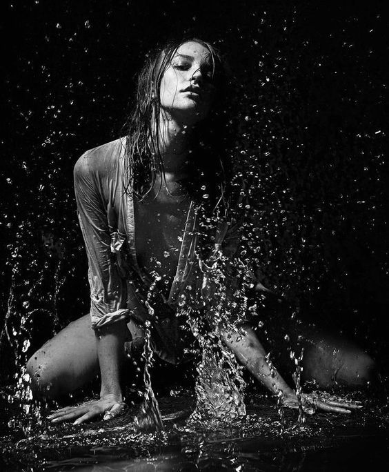 the image is a dramatic, black and white portrait of a young woman. she is sitting in water, with splashes and droplets surrounding her.  her long, wet hair frames her face, and she appears to be looking directly at the camera with a serene yet slightly mysterious expression. she is wearing a wet, open shirt. the overall mood is sensual, mysterious, and artistic, with a strong emphasis on contrast and dramatic lighting. the background is dark, drawing focus entirely to the woman in the center.