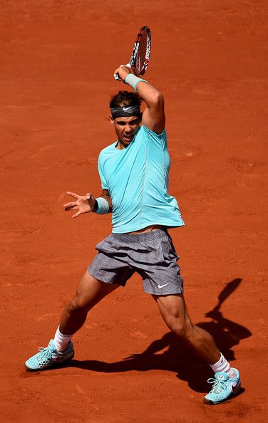 a male tennis player in a blue shirt and grey shorts  is playing on a red clay court. he is holding a tennis racquet in his right hand and is in midswing. his left leg is bent, and his right leg is straight out. his left hand is up in the air, and he is looking at the ball. he is wearing a headband and has a determined expression on his face. the image captures the action and skill of a professional tennis player.