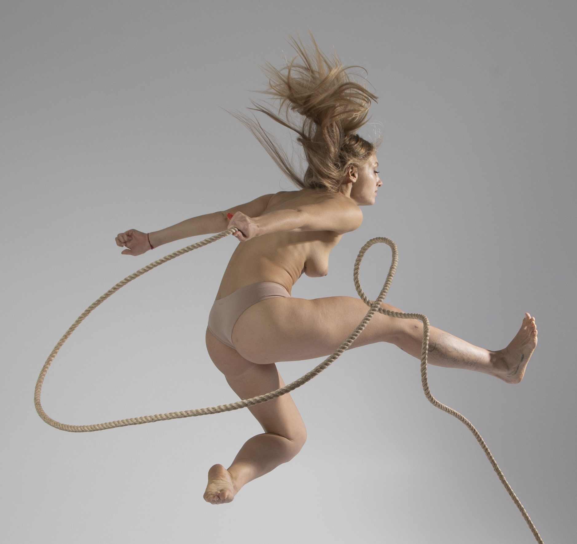 a nude blonde woman is jumping in the air while holding a rope. she is wearing beige underwear. the background is a plain light gray color. the woman's hair is flying upwards, suggesting movement.