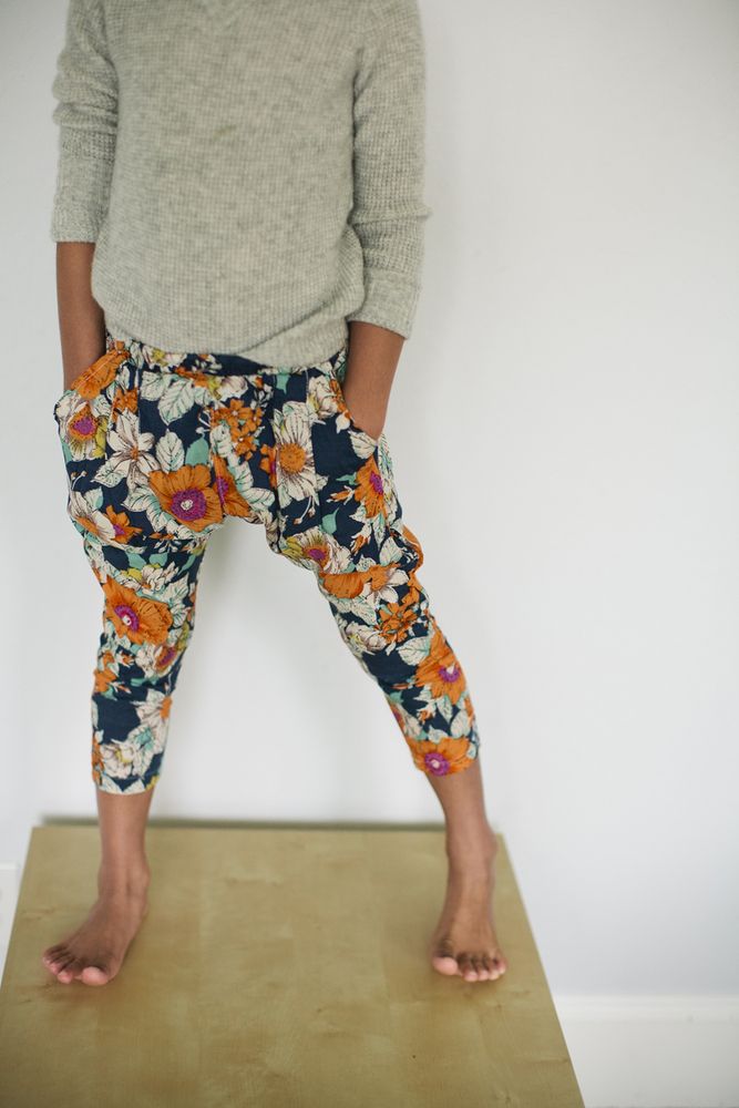 a young girl with light skin is wearing a light grey sweater and colorful pants. the pants are blue with orange and pink flowers. she is standing barefoot on a wooden floor in front of a white wall.  she is looking down,  and  her arms are relaxed.  her hair is not visible.  her hands are in her pockets.