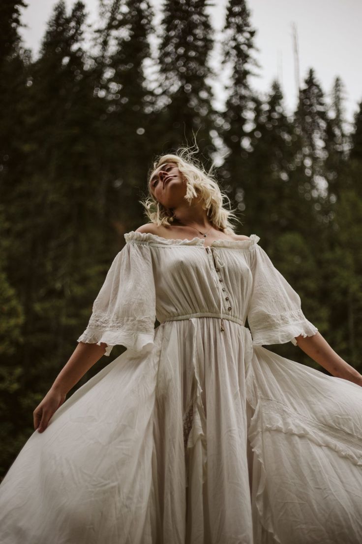 a woman in a white offtheshoulder dress stands in a forest with her arms outstretched. she has blonde hair and is looking up towards the sky. the background is filled with trees. it's daytime and the scene is set in nature.