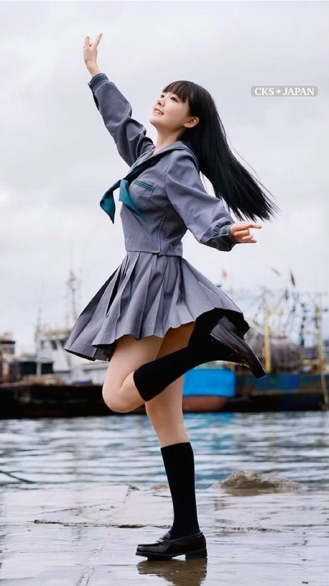 a young asian woman in a gray sailorstyle school uniform is captured midleap on a harbor dock. her arms are outstretched, her head tilted back, and her skirt billows around her, conveying a sense of joy and freedom. the overcast sky and calm harbor water create a muted backdrop, emphasizing the energy of her movement.