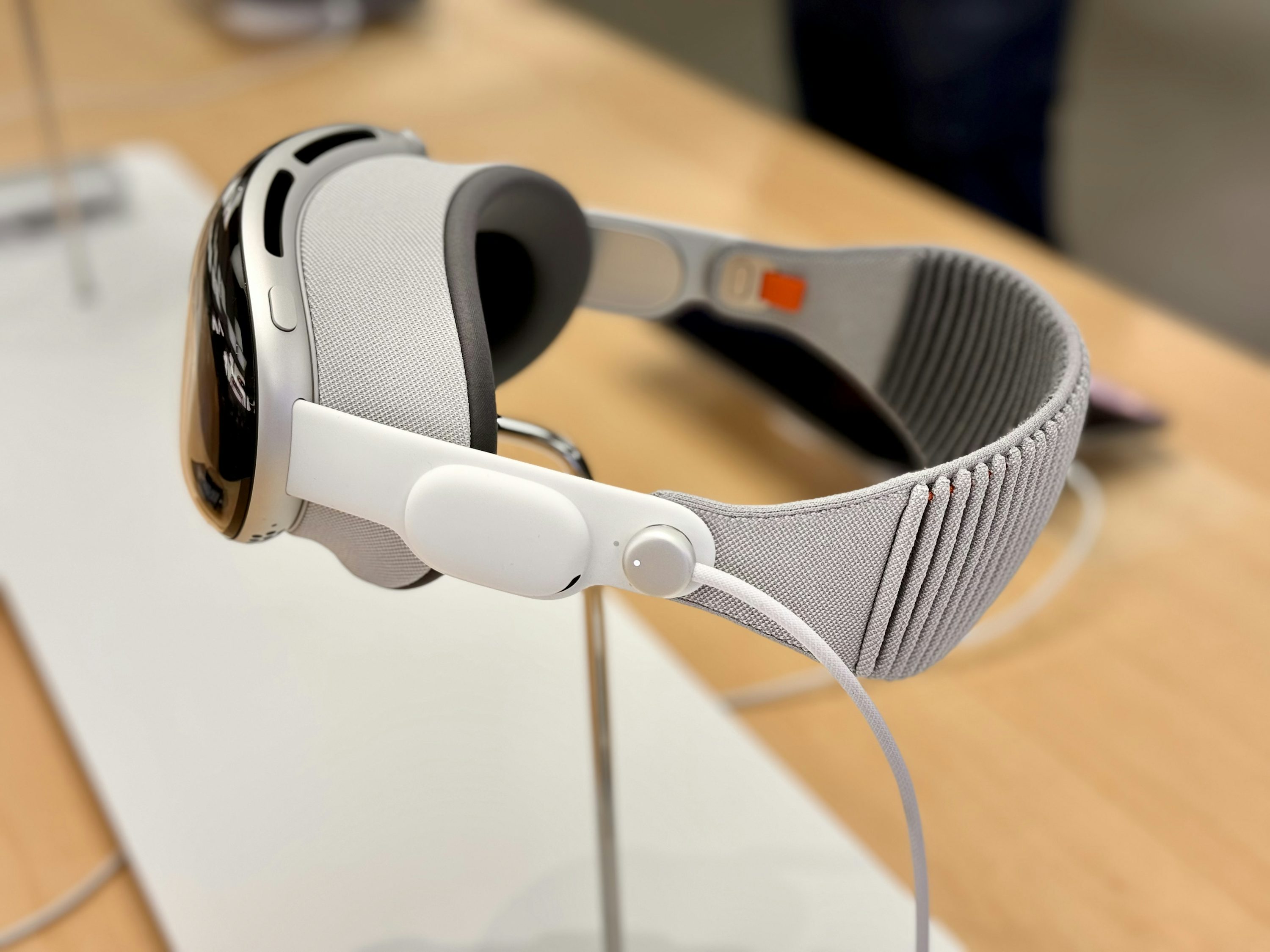 the image shows the apple vision pro headset, a mixed reality device, on a display stand. the headset has a sleek, futuristic design with a glossy black front, a gray fabric headband, and white accents. a cable is attached to the side of the headset. the background is a blurry, lightcolored surface.