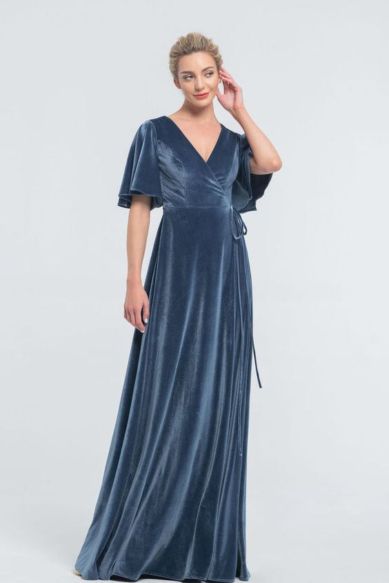 a woman in a long blue velvet dress with a vneck and flutter sleeves stands in a studio setting. she has her hand on her face and is looking to the side.