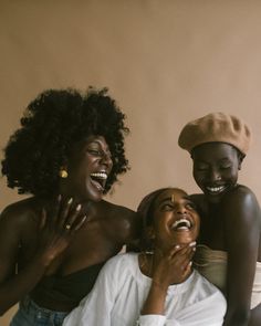 three beautiful black women are captured in a moment of pure joy, their laughter and radiant smiles illuminating the image. they stand close together, their arms intertwined, showcasing a powerful sense of friendship and connection. the warm, soft lighting enhances the intimacy of the scene, emphasizing the genuine happiness they exude.  their stylish clothing and the neutral backdrop further contribute to the overall aesthetic of the image, highlighting the beauty and strength of these women.