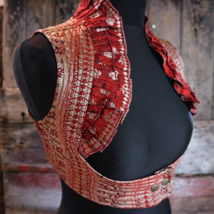 a red and gold brocade vest with a ruffled collar is displayed on a black mannequin against a rustic wooden backdrop. the vest features intricate patterns and is fastened with two buttons at the back.