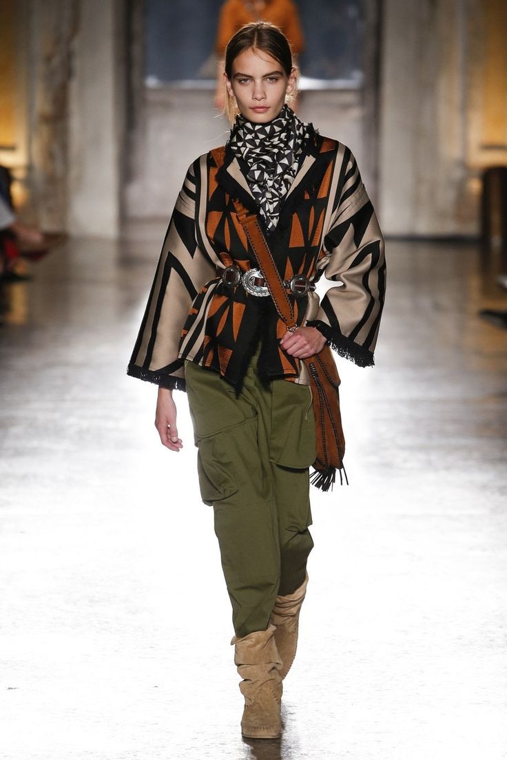 a fashion model walks the runway wearing a stylish ensemble. the outfit consists of a patterned poncho with fringed edges, olive green cargo pants, tan suede boots, a black and white patterned scarf, and a brown leather belt with a large buckle. she also carries a fringed brown leather handbag. the model has her hair tied back in a low bun and minimal makeup. the background is a blurred out runway setting. the overall style is a blend of bohemian and western influences.