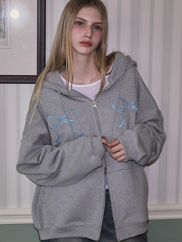a young woman with blond hair is wearing a gray zipup hoodie with light blue ribbon embroidery on the chest. she is standing indoors against a striped wall. the hoodie is slightly oversized and she has a white shirt underneath. her expression is neutral. there is a framed picture on the wall to her left.