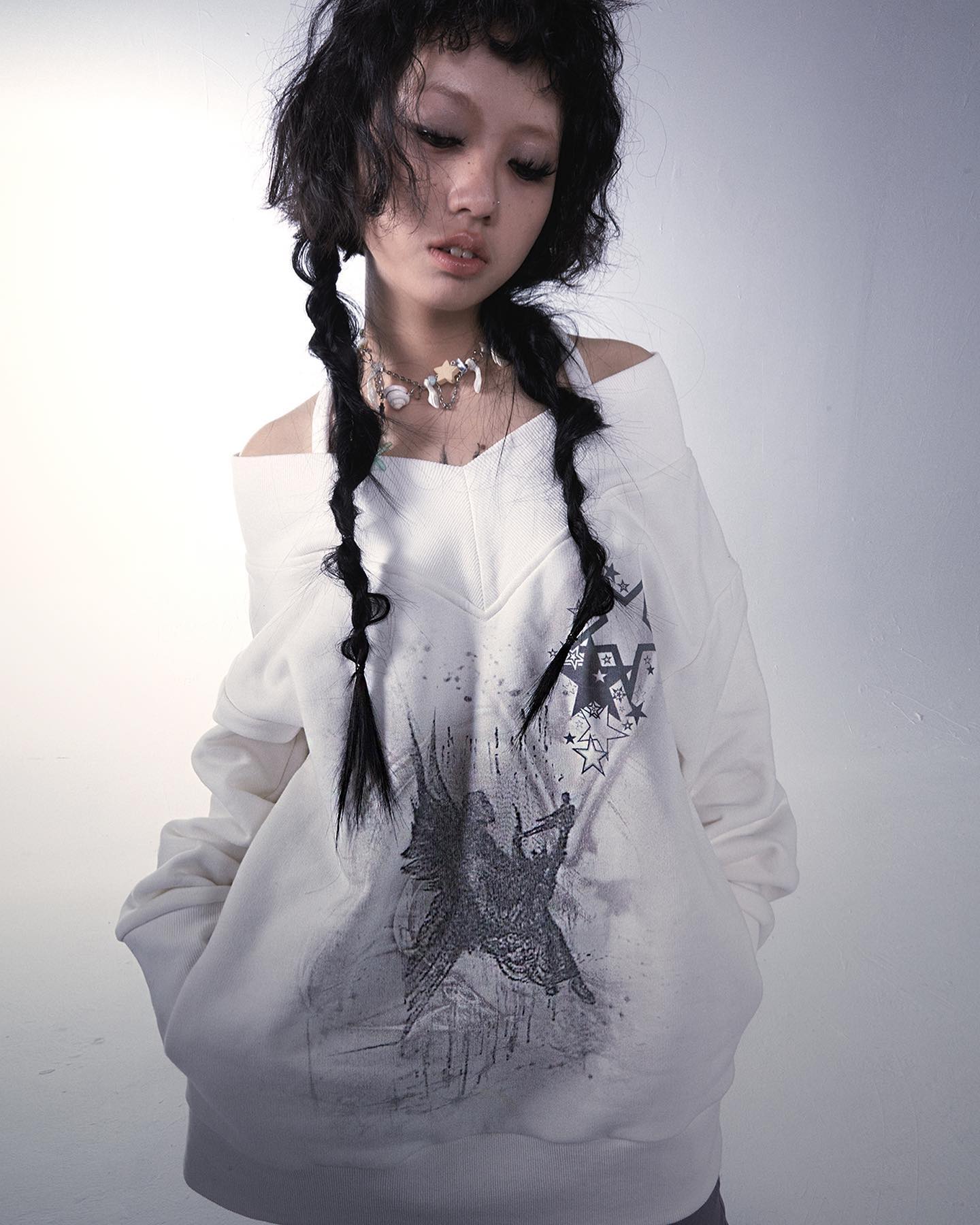 a young asian woman with long black hair styled in pigtails models a white offtheshoulder sweatshirt with a graphic print of an angel and stars. the sweatshirt has a vneck and kangaroo pocket. she wears a delicate necklace. the background is a plain light gray. the overall style is grunge and alternative.