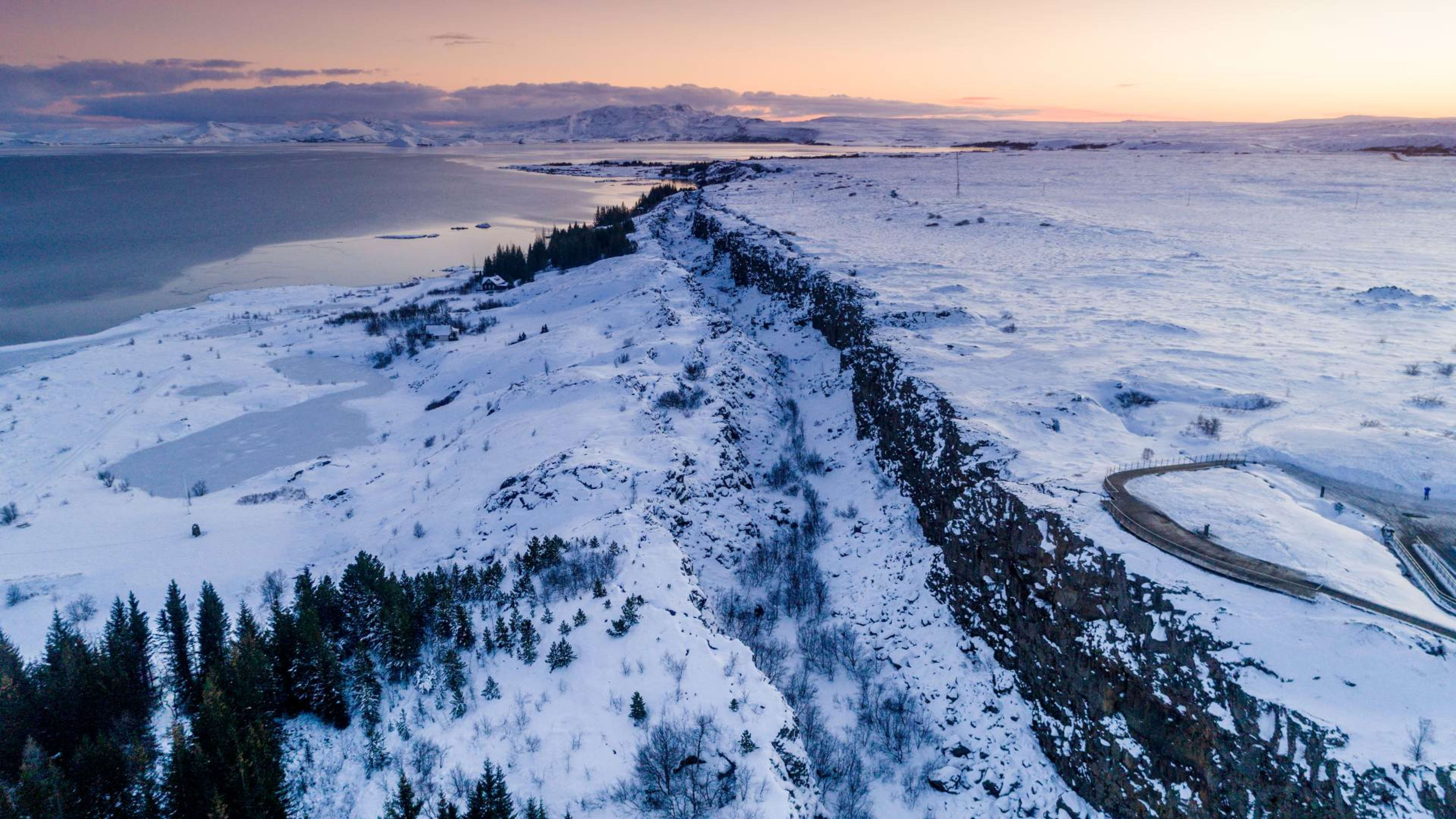 a breathtaking aerial view of a snowcovered landscape in iceland, featuring a dramatic cliff that cuts through the pristine white landscape. the frozen lake in the foreground reflects the soft pink hues of the setting sun, casting a magical glow over the scene. the rugged cliffs,  the glistening ice and the soft light of the sky create a truly mesmerizing sight.
