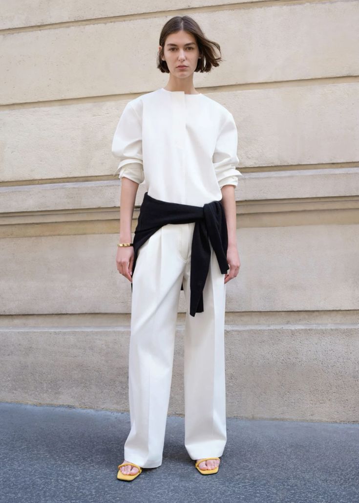 a woman standing in front of a white wall wearing a white shirt, black sweater as a belt, and white pants, yellow sandals. she is looking at the camera.  the background is a white wall with a rough texture.