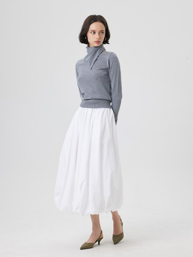 a woman stands in a studio, wearing a gray turtleneck sweater and a white balloon skirt. she is also wearing olive green heels. the image is a full body shot of the woman.