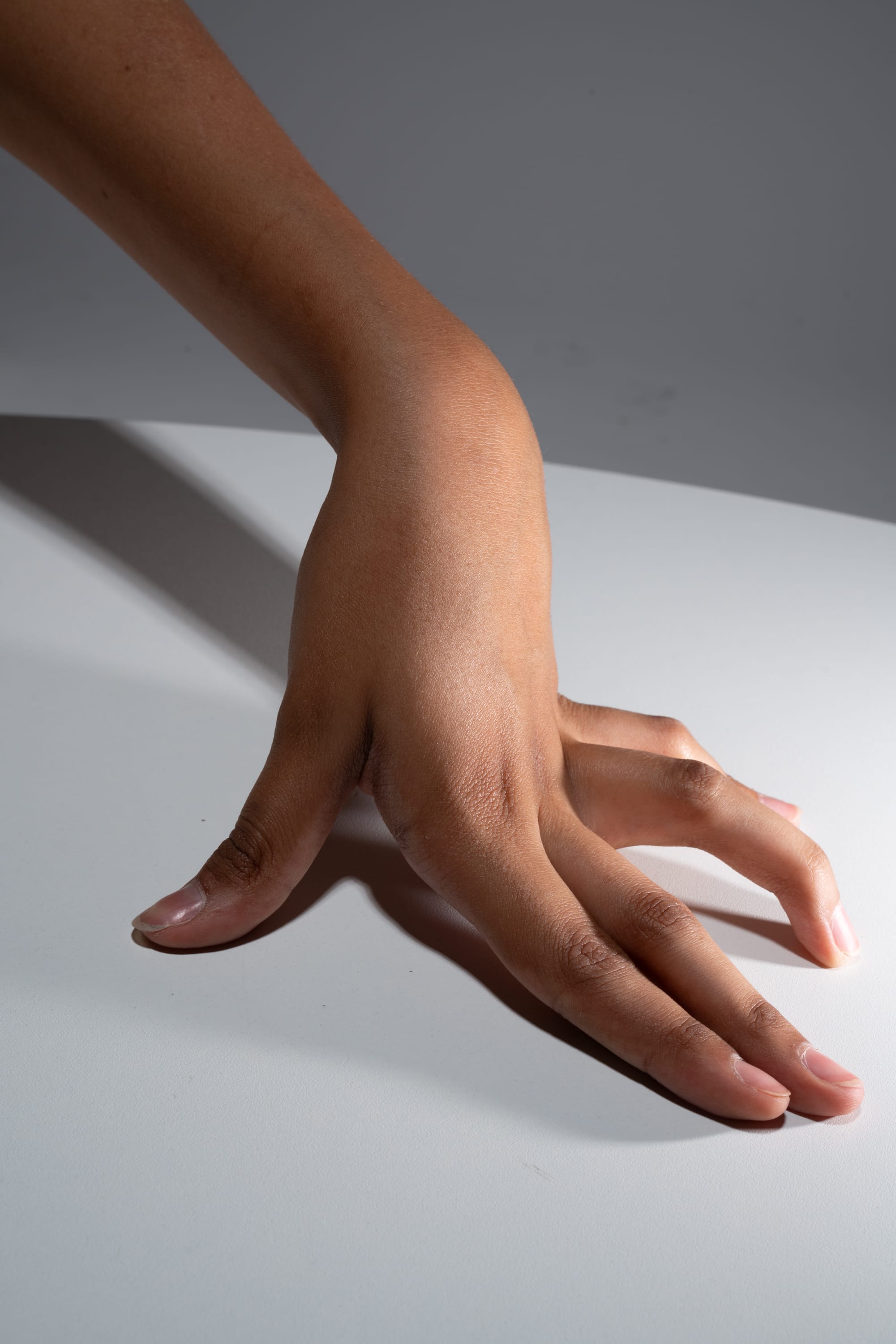 a closeup image of a hand with dark skin tone resting on a white table. the image is lit by a studio light, creating a sharp contrast between the hand and the background.