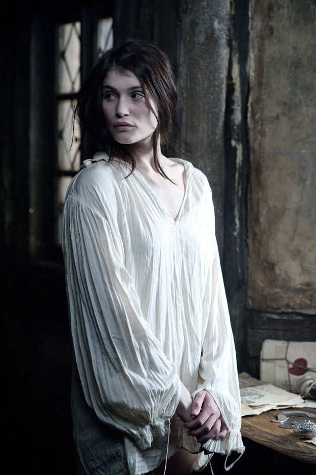 a young woman with dark hair, wearing a loose white shirt, stands in a dimly lit, rustic interior. she is positioned near a window, which provides a soft, natural light source. the overall color palette is muted, with shades of brown, gray, and white dominating the scene.