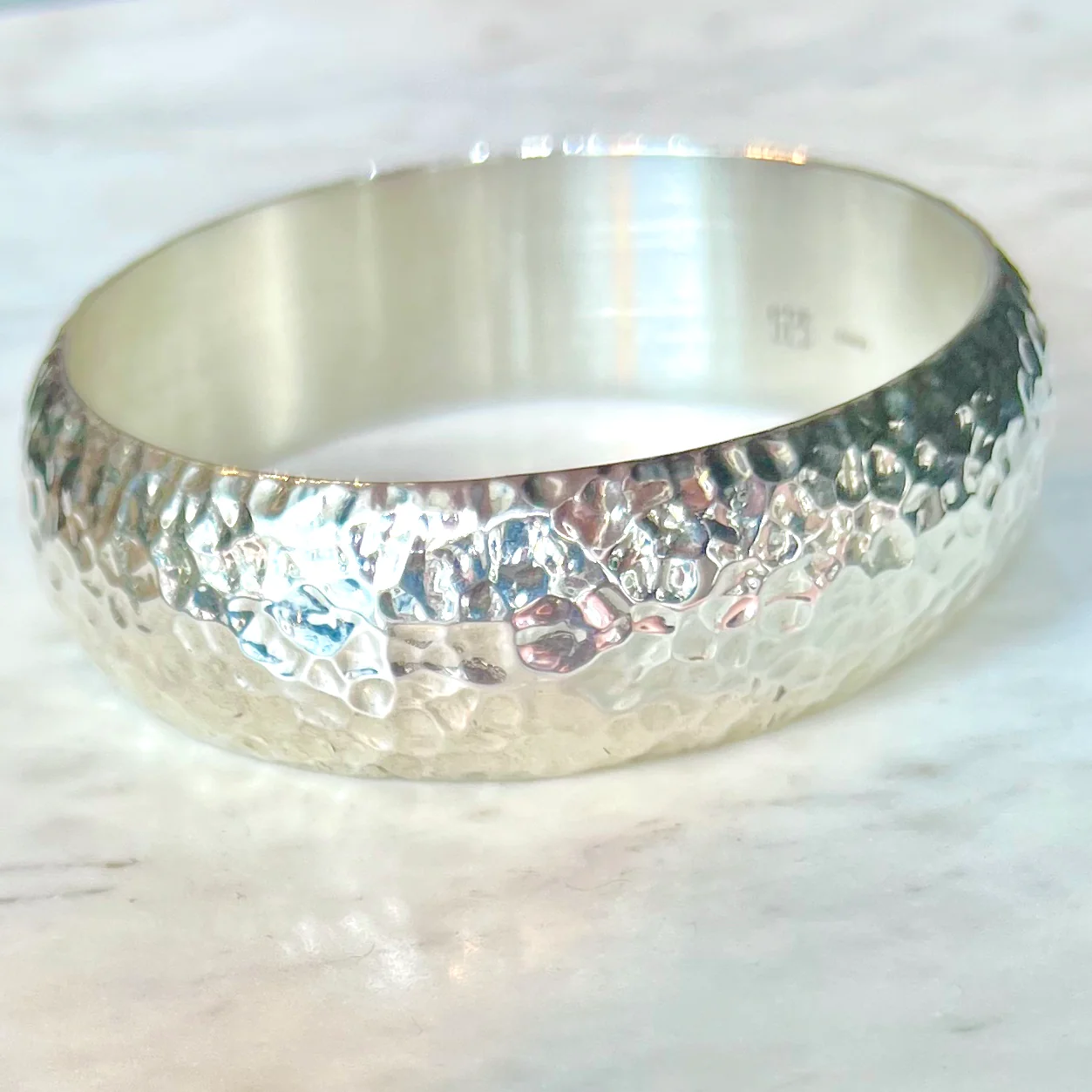 this is an image of a silver hammered bracelet sitting on a white marble surface. the bracelet is wide and has a textured surface with a hammered design. the bracelet is made of sterling silver, as indicated by the "925" hallmark visible on the inside. the image is lit with natural light.