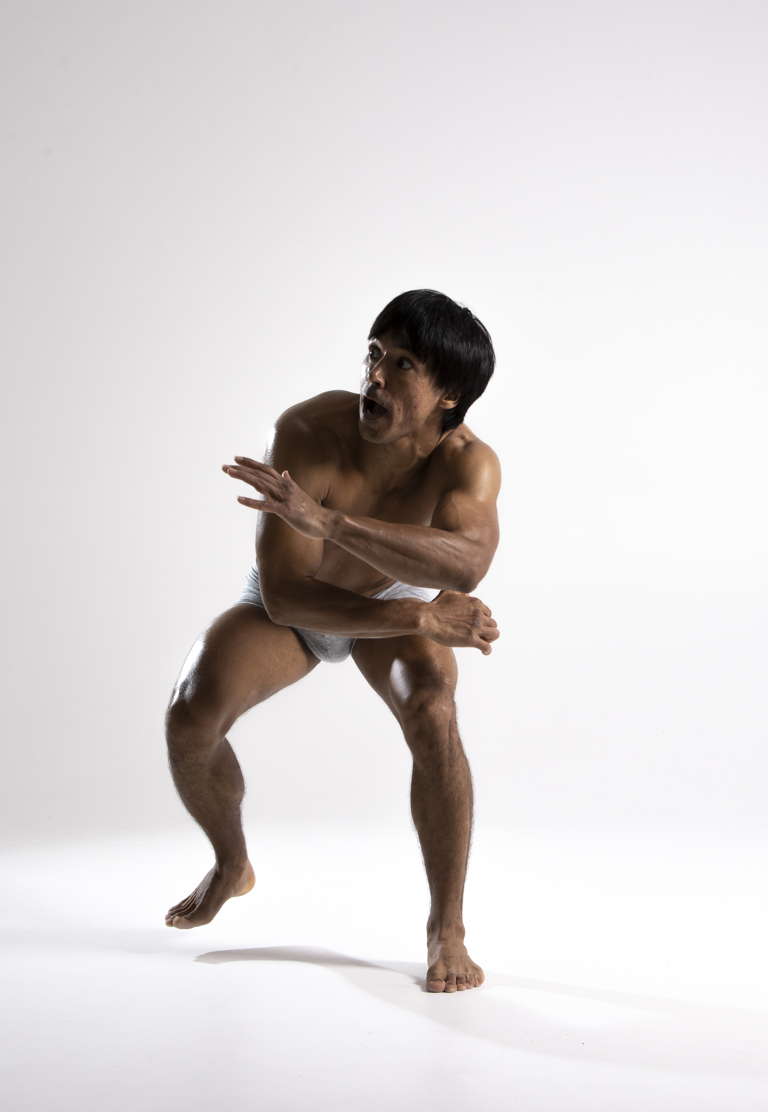 a studio shot depicts a muscular asian man wearing underwear. he is posed with his arms crossed in front of him, his body angled with one foot slightly raised off the ground. his expression is one of fear and surprise, and he is looking up and away from the viewer.