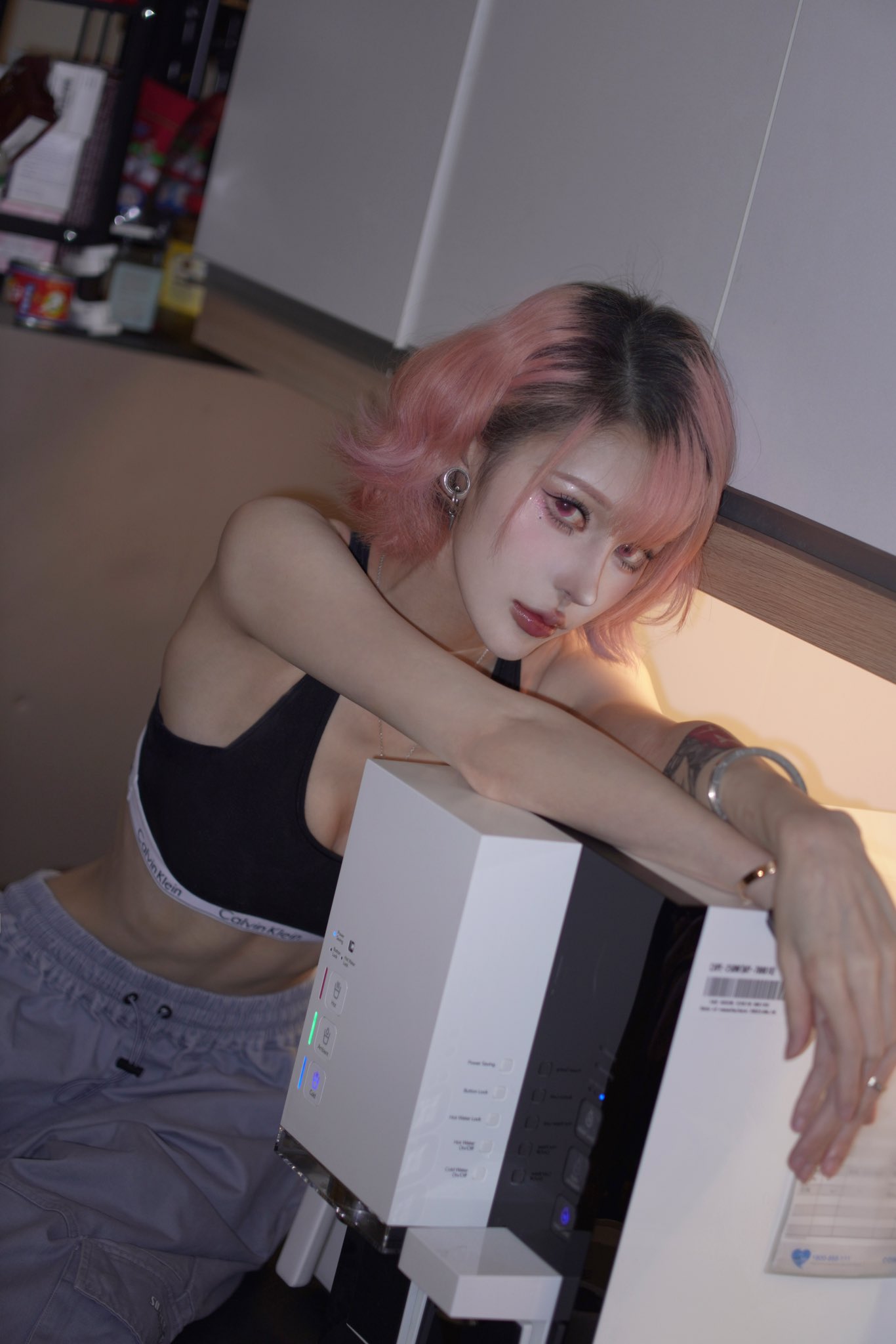 a young woman with short, pink hair is posing in a kitchen. she is wearing a black sports bra and grey sweatpants. she has makeup on and is wearing jewelry, including earrings and a bracelet. she is leaning on a white appliance, possibly a water dispenser. the overall aesthetic is stylish and somewhat edgy.