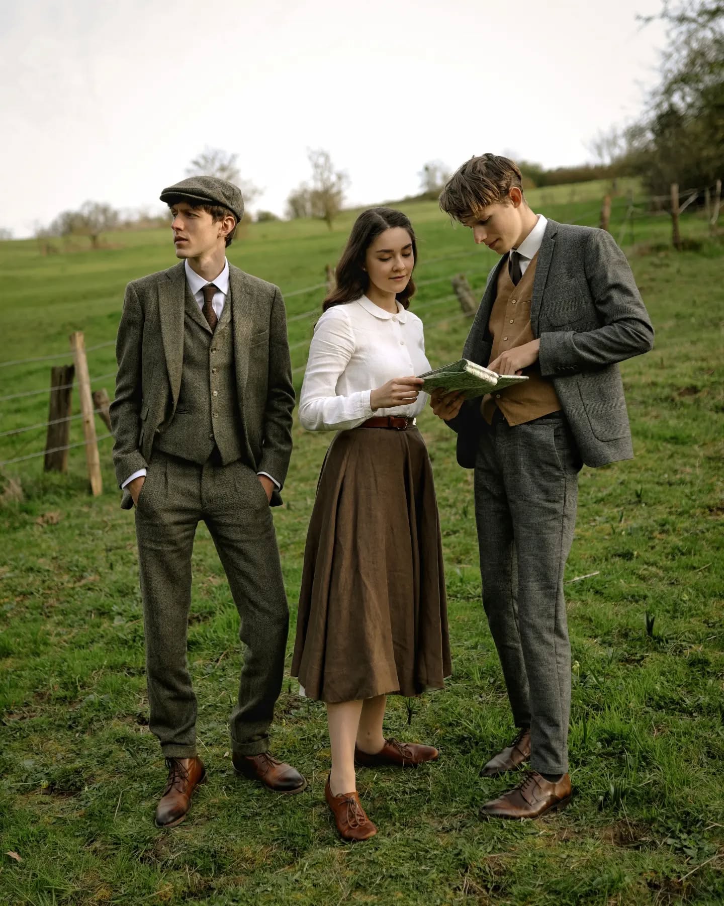 hree young adults, two men and one woman, are standing in a green field in the countryside. the men are wearing vintagestyle tweed suits, and the woman is wearing a brown skirt and a white blouse. they appear to be looking at a book or map together. the scene is set outdoors in a natural, rural setting, suggesting a relaxed and friendly atmosphere. the clothing style hints at a 1920s aesthetic.