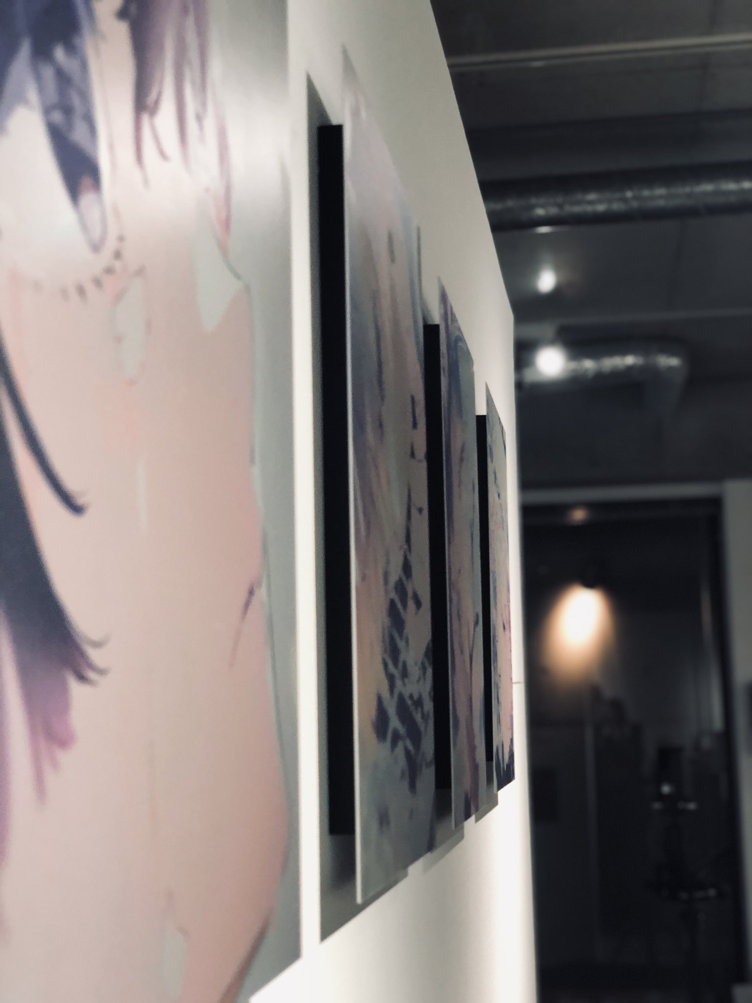the image shows a series of abstract paintings hanging on a white wall in an art gallery. the paintings are mounted on black frames and are illuminated by spotlights. the gallery has a modern, industrial feel with exposed ceilings and concrete floors. the perspective is from the side, looking down a hallway lined with paintings.