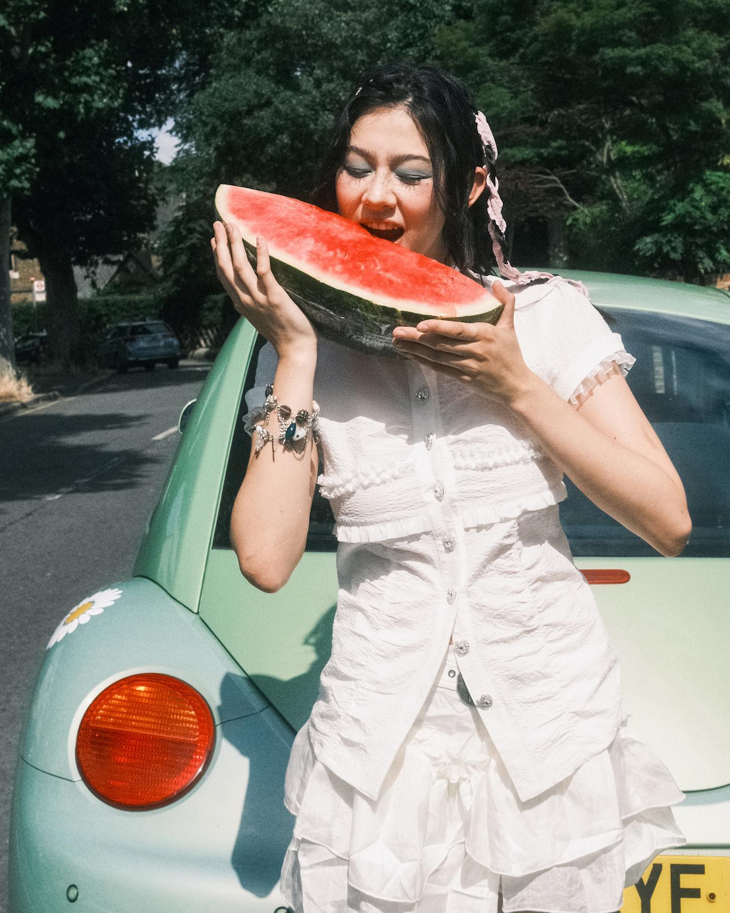 a young woman in a white ruffled dress is standing next to a vintage green volkswagen beetle. she is happily biting into a large slice of watermelon. the scene is set on a sunny street, with trees and other cars in the background. the overall aesthetic is summery and cheerful.