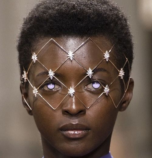a closeup portrait of a black model with short black hair on the runway, she's wearing a diamond starshaped headpiece covering her eyes. the model's face is covered with a decorative wire mesh structure, adorned with multiple diamond starshaped embellishments. her eyes are a striking shade of purple, adding an ethereal touch to the overall look. the model is confidently looking forward, showcasing her fierce and glamorous style.