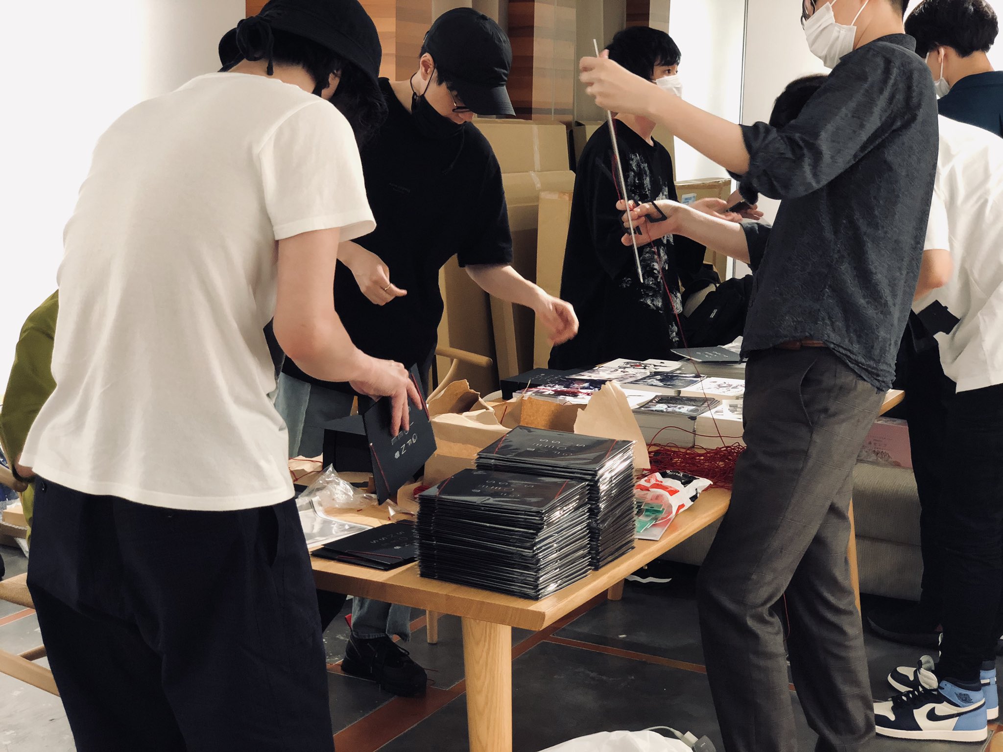 a group of young men are working together indoors, packaging what appears to be music albums or cds.  they are surrounded by boxes, packaging materials, and finished products on a wooden table. the scene suggests a busy and productive atmosphere, possibly behind the scenes at a music label or distribution center.