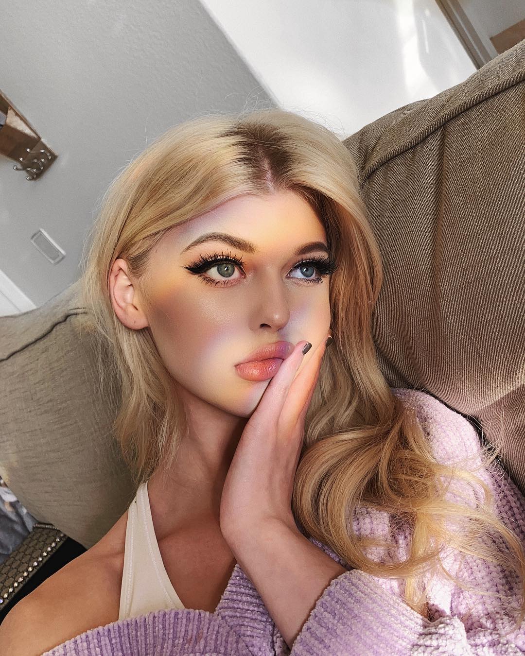 a portrait of a blonde woman with heavy eye makeup wearing a pink sweater and touching her face. she is sitting on a couch and a beam of light is shining across her face.