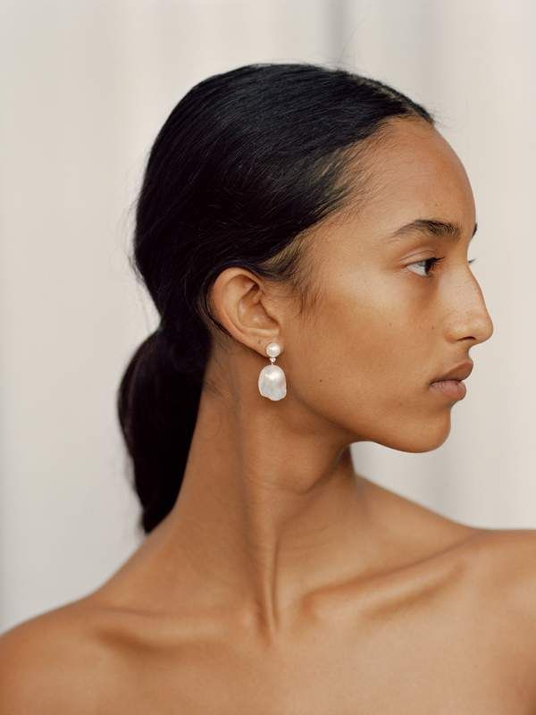 a woman with long dark hair is looking off to the right side of the frame.  she is wearing a pair of pearl earrings. the background is a soft white.  her skin is smooth and natural.