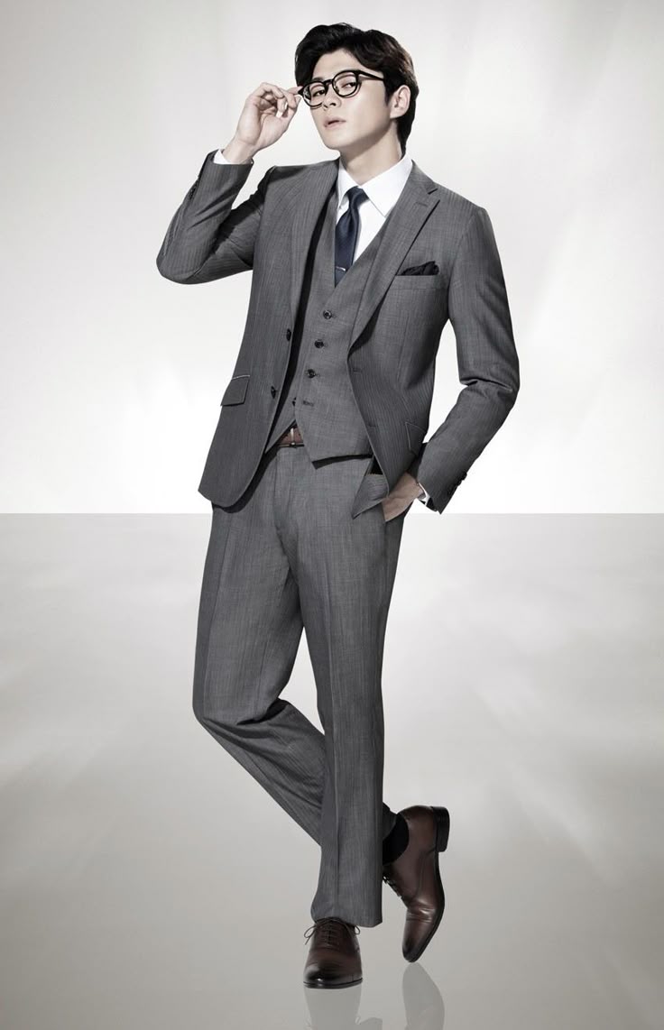 a man in a grey threepiece suit and glasses stands confidently with one hand raised near his glasses and the other in his pocket. he's wearing brown dress shoes and a tie. the background is a light gradient.