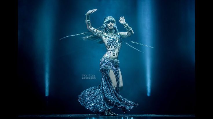 a woman is belly dancing on a stage with a blue spotlight on her. she is wearing a traditional costume with intricate jewelry and a headpiece. the dress is long and flowing, with a slit up the side. she has bracelets on her wrists and is holding her hands up in the air.
