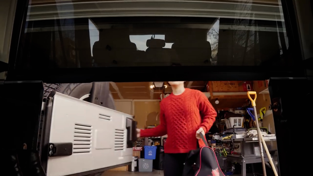 a woman in a red sweater is standing in a garage, facing the open back door of a car.  she appears to be getting ready to either load or unload items from the vehicle.  the garage is cluttered with tools and other items.