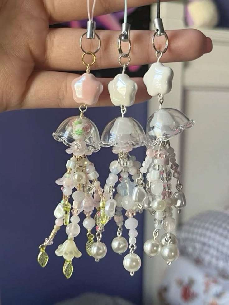 a hand holds three handmade jellyfishshaped cellphone charms. each charm has a starshaped bead, a translucent, bellshaped bead, and multiple beaded tassels hanging from the bell. the charm on the left has a light pink star and a bell filled with pink and green floral elements. the center charm has a white star. the charm on the right is also white. the beaded tassels are strung with pearls, light pink and white beads, and leafshaped accents.