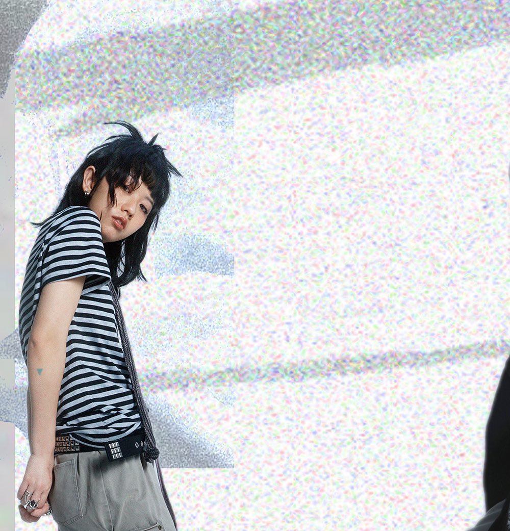 the image features a young woman, possibly a fashion model, posing against a heavily textured, noisy background with hints of pastel colors. she is wearing a horizontally striped black and white tshirt, olive green cargo pants, and a studded belt.  she has short, dark hair styled somewhat messily.  she's wearing rings and appears to have a dark tie or scarf draped loosely around her neck. her pose is slightly turned away from the camera, with her gaze directed over her shoulder. the overall style is edgy and grungy, with a focus on fashion and style.