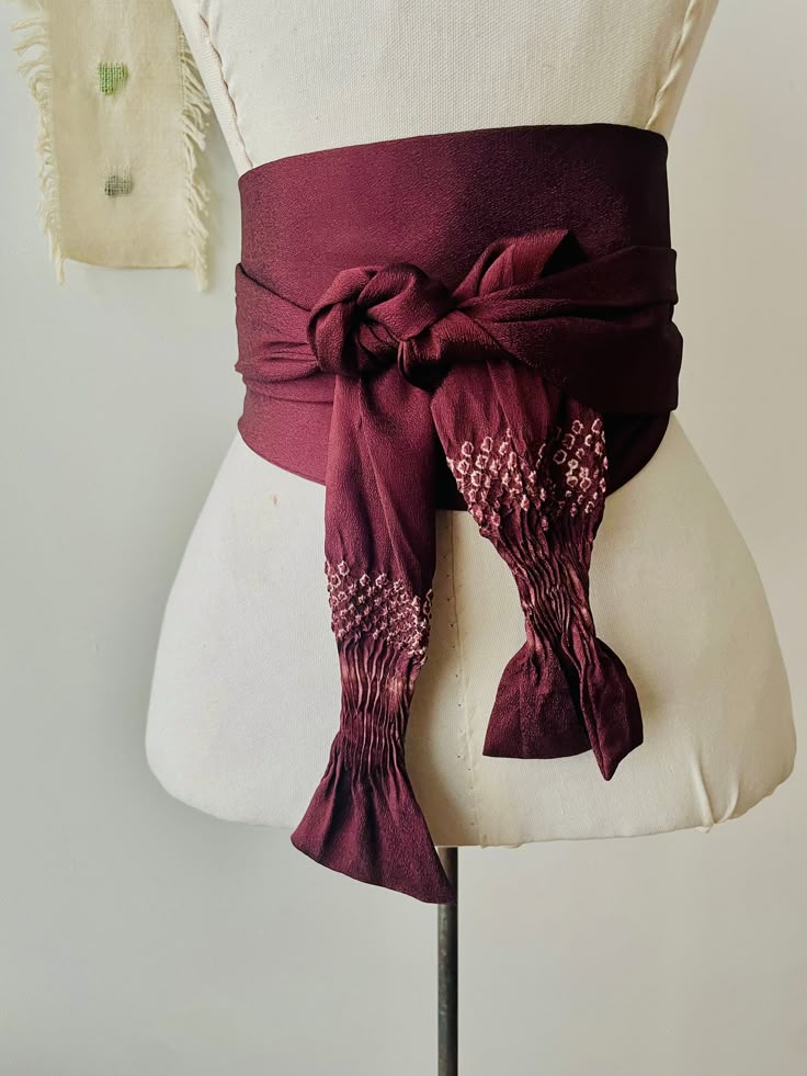 a burgundy obi belt is tied around a mannequin in the image. the belt is made of fabric and has a knot. the mannequin is white.