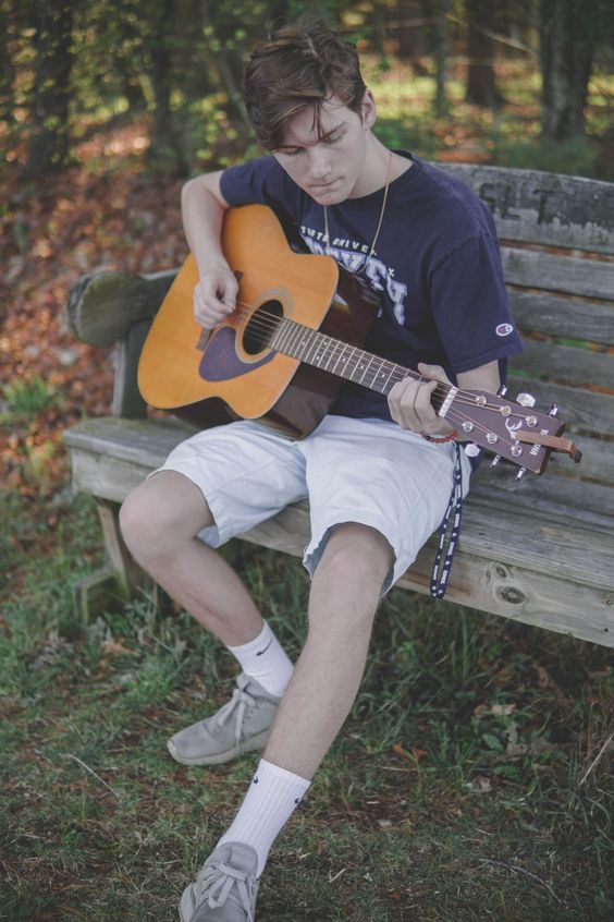 a young man sits on a wooden bench outdoors playing a guitar. he is wearing white shorts, a blue shirt, and white sneakers. the man is smiling, appears relaxed, and seems to be enjoying playing his guitar.  there are trees and green grass in the background, suggesting a park or outdoor setting.
