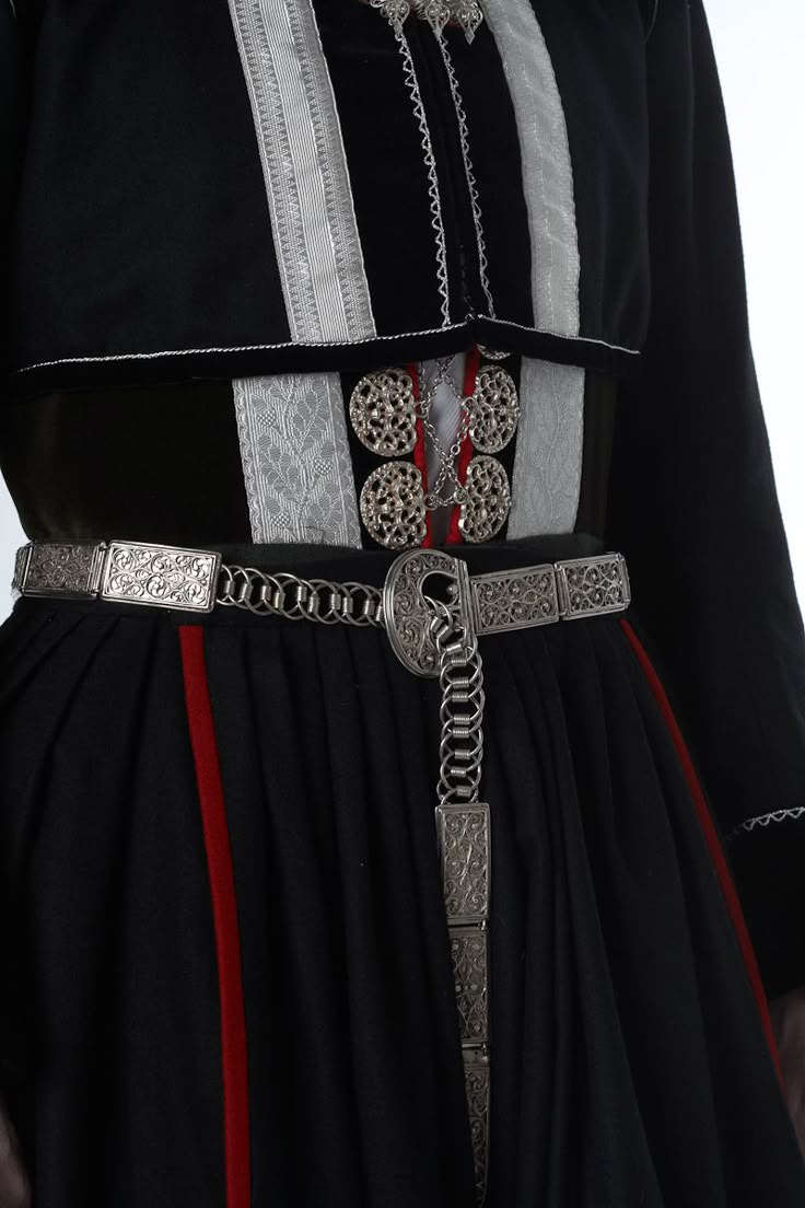 a closeup shot of a traditional folk costume, predominantly black with silver ornamental details. the outfit features a black dress or skirt with a vertical red stripe. a silver belt cinches the waist, featuring an intricate design with decorative panels connected by chains. the bodice is adorned with silver embroidery and ornate metal fastenings, adding to the garment's rich, decorative appeal. the overall impression is one of cultural heritage and formal elegance.