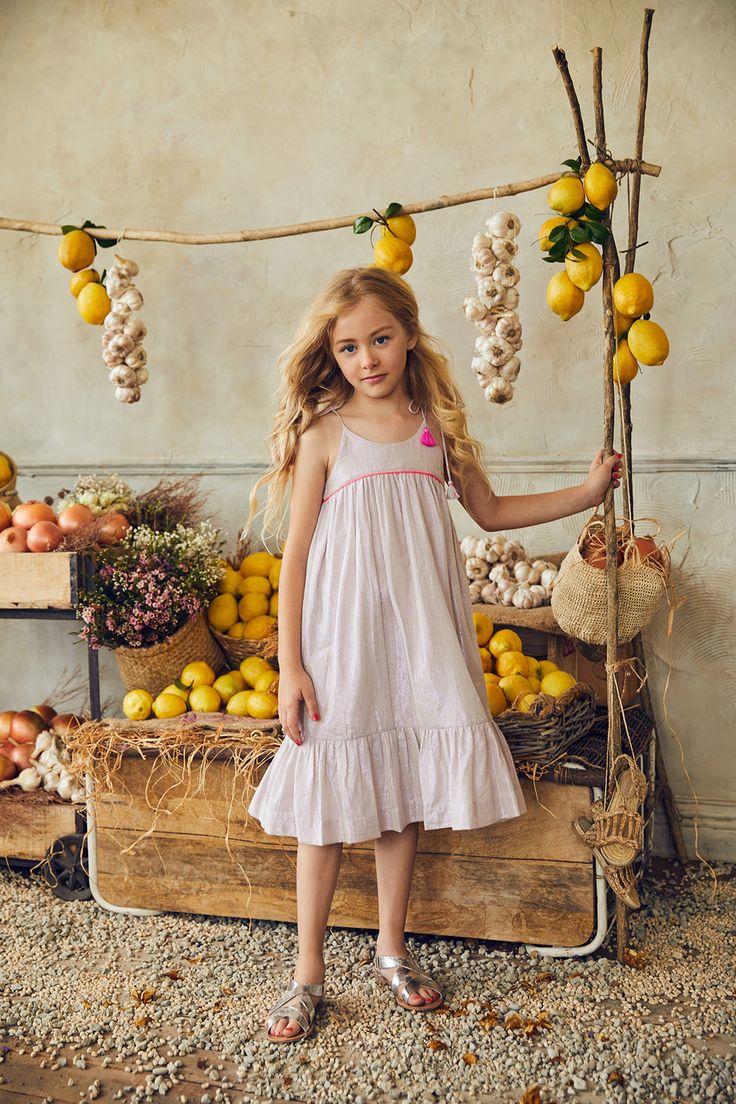 a young blonde girl in a light purple summer dress stands in front of a rustic wooden market stall laden with lemons and garlic. she is smiling slightly and looking at the camera. the overall aesthetic is rustic, summery, and charming. the girl's dress has a ruffled hem and thin straps. she is wearing silver sandals.  the scene is brightly lit with natural light, giving it a warm and inviting feel.