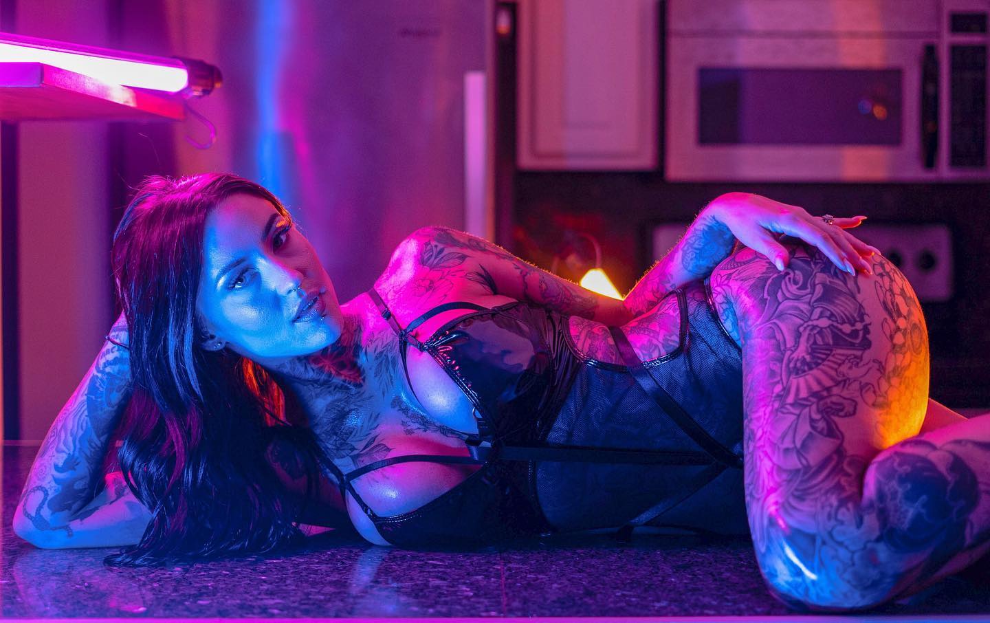 the image is a stylized portrait of a heavily tattooed woman lying on a kitchen counter.  she's wearing black lingerie with a mesh overlay, and the lighting is dramatic, using strong neon purple and blue hues that accentuate her tattoos and the textures of her clothing. the woman's pose is sensual, and she looks directly at the camera with a confident expression. the kitchen background is blurred, drawing focus to the model. the overall atmosphere is dark, moody, and glamorous.
