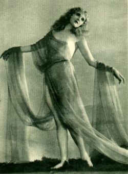 a black and white photograph of a woman, possibly a dancer, in a flowing, sheer gown. she's posed gracefully with a pensive expression. the image has a vintage, artistic quality, possibly from the 1920s, evoking a sense of ethereal beauty and mystique. the background is blurred, focusing attention on the woman and her elegant pose. the fabric of her dress drapes beautifully around her body.