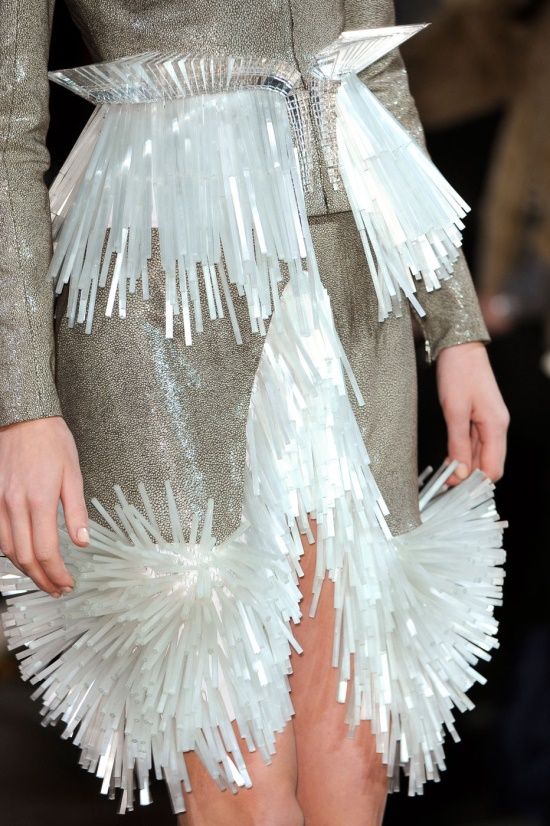this is a photo of a woman wearing a silver and white dress with a fringe skirt. the dress is made of a delicate, silky fabric and the fringe is made of white, plastic material. the model is standing on a runway, posing for the camera. the dress is very elegant and sophisticated, and the fringe adds a touch of whimsy to the outfit.  the fashion is a mix of modern and vintage design.