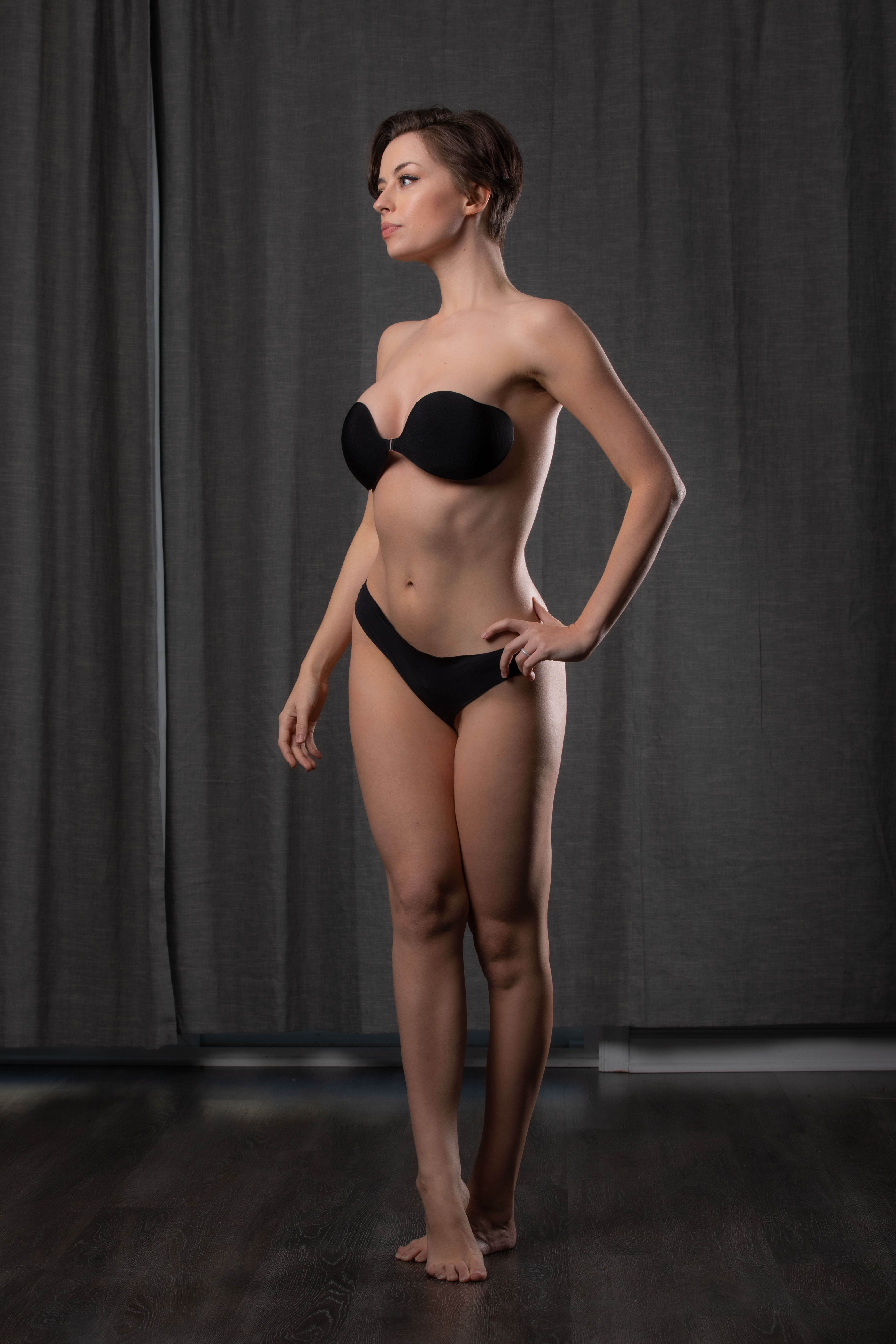 a woman with short hair is standing in a studio setting. she is wearing black underwear and a black bra. she is posing in front of a gray background with bare feet.