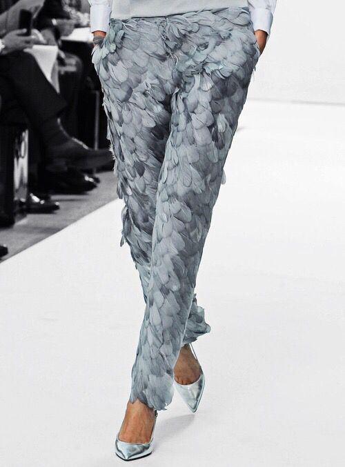 a model wearing grey pants made of feathers and silver high heels walks down a white runway. the background is blurred and shows other people.