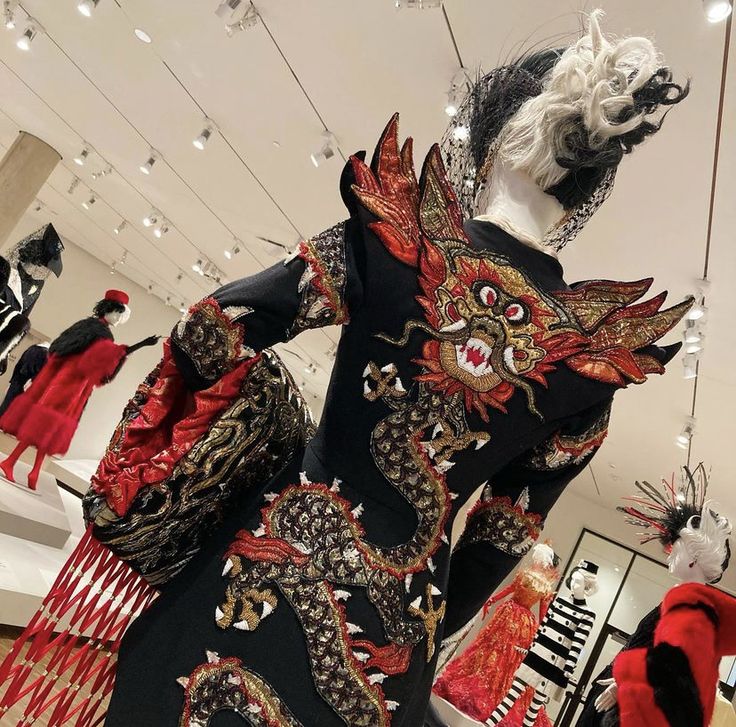 this image showcases a mannequin wearing a striking black dress adorned with intricate dragon embroidery in red and gold. the dress is displayed in what appears to be a museum or fashion exhibit, alongside other mannequins dressed in various styles. the mannequin is also wearing a unique headdress. the setting is welllit and modern.