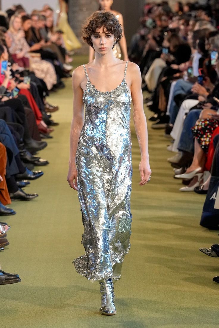 a model walks down a runway in a fashion show. she is wearing a silver sequin dress and silver boots. the audience is seated on either side of the runway.