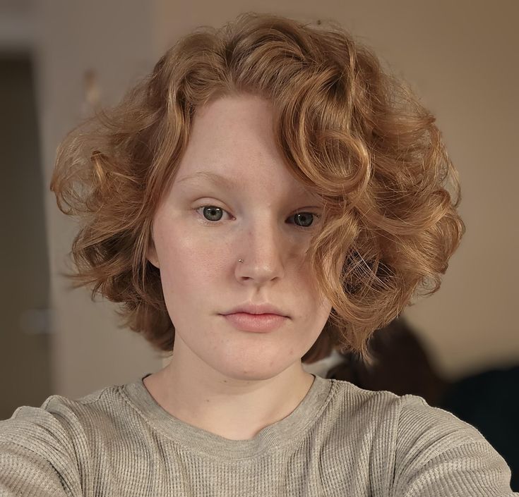 the image is a closeup portrait of a fairskinned woman with curly red hair. she is wearing a gray waffleknit shirt and has a nose piercing. the background is a neutral color, and the lighting appears to be natural. she appears to be taking a selfie.