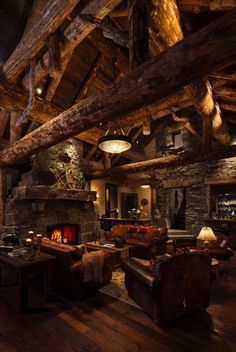 the image shows a rustic living room in a log cabin style. the room features a large stone fireplace, exposed wooden logs on the ceiling, and comfortable furniture including armchairs and a sofa. the lighting is warm and inviting, with a lamp and overhead lighting creating a cozy atmosphere.