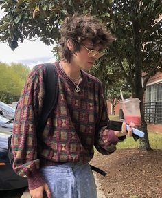 a young man in a trendy checkered sweater, sunglasses, and jeans holds an iced beverage. he stands outdoors, possibly on a sidewalk, in the daytime.